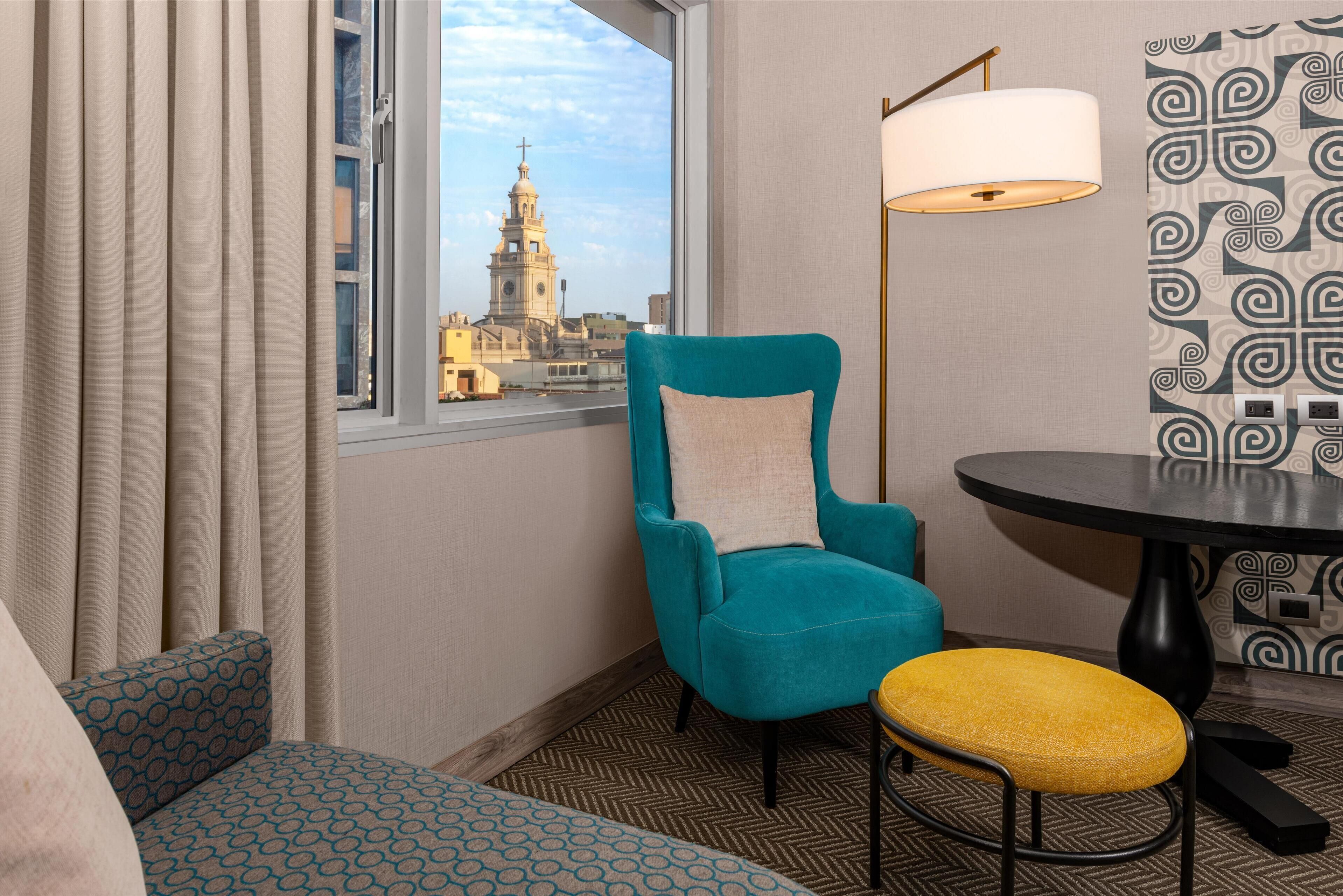 A hotel room with a teal armchair, a yellow stool, a round table, and a view of a building with a bell tower.