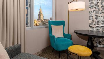 A hotel room with a teal armchair, a yellow stool, a round table, and a view of a building with a bell tower.