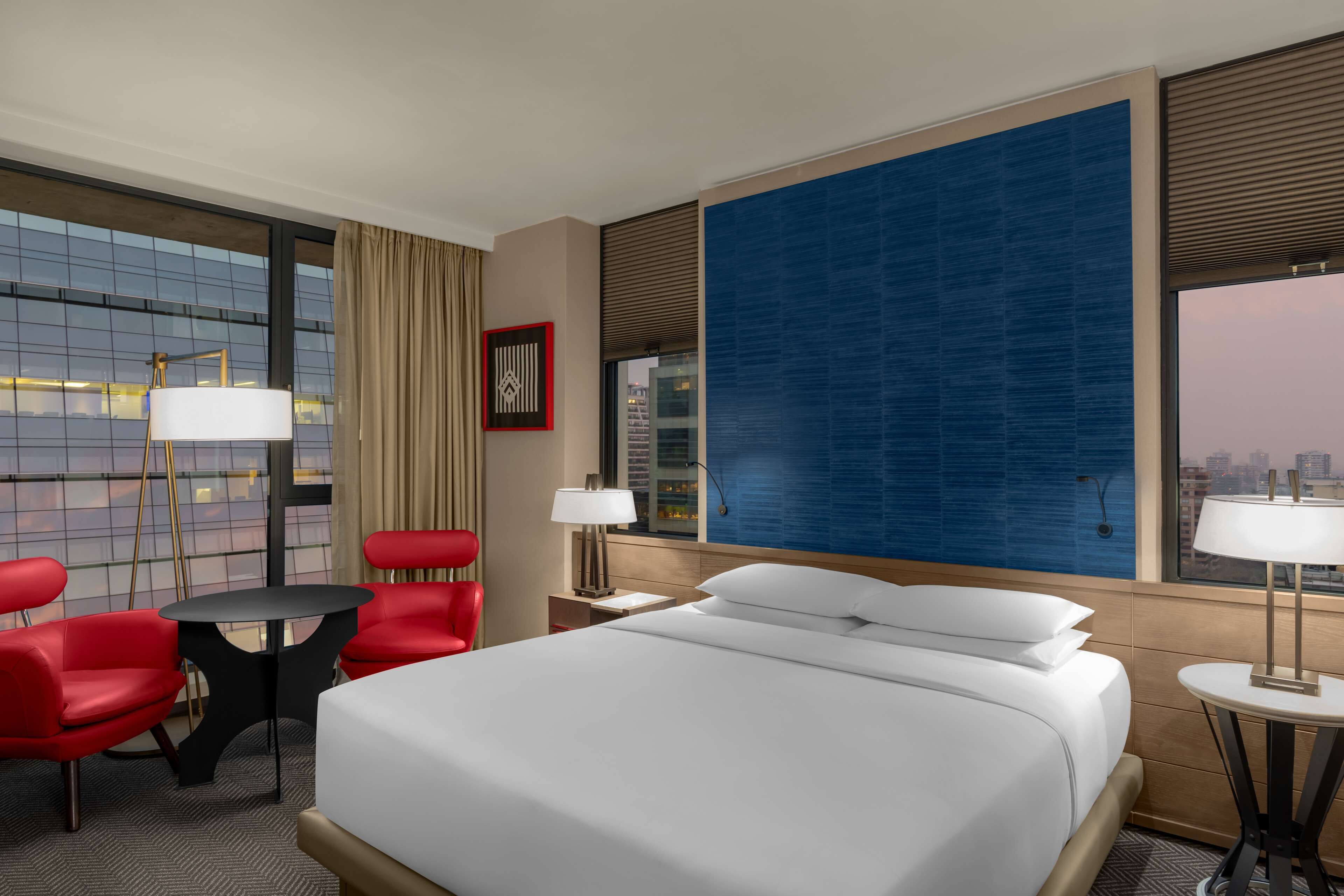 Habitación de hotel con una cama grande, sillas rojas, una mesita y un ventanal con vistas a la ciudad.