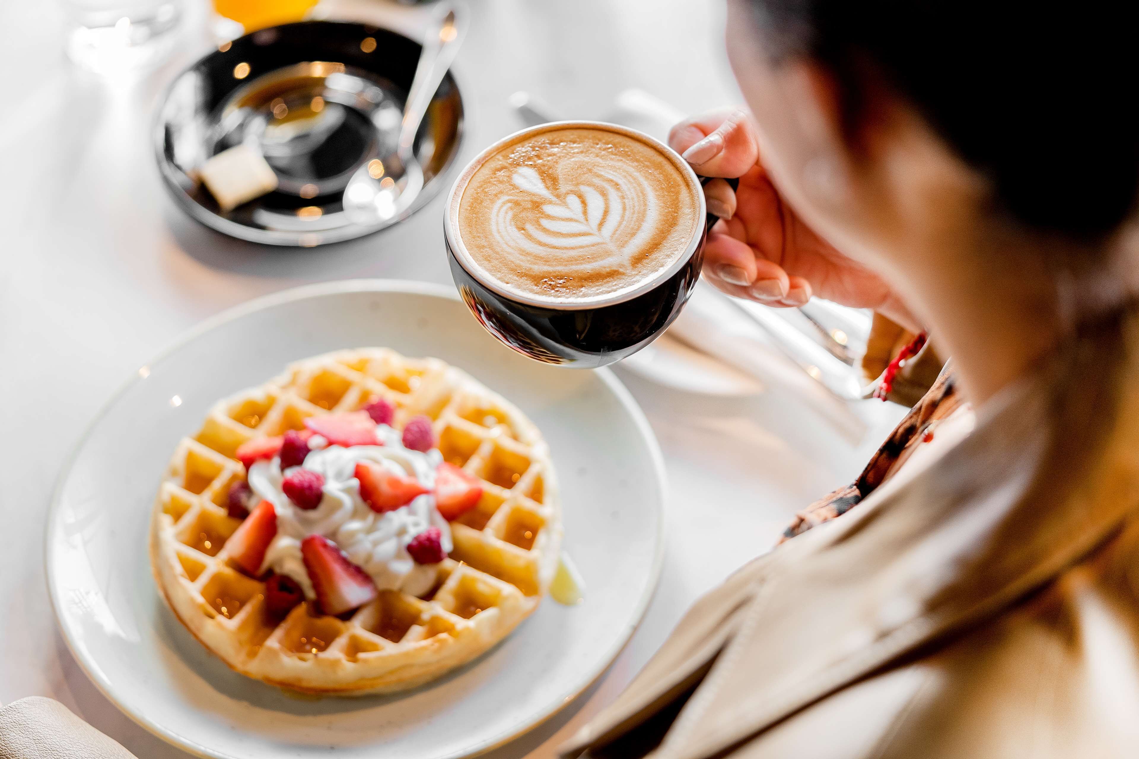 A person holding a cup of coffee with latte art, a waffle topped with whipped cream and fruit, and a small bowl of syrup on a table.