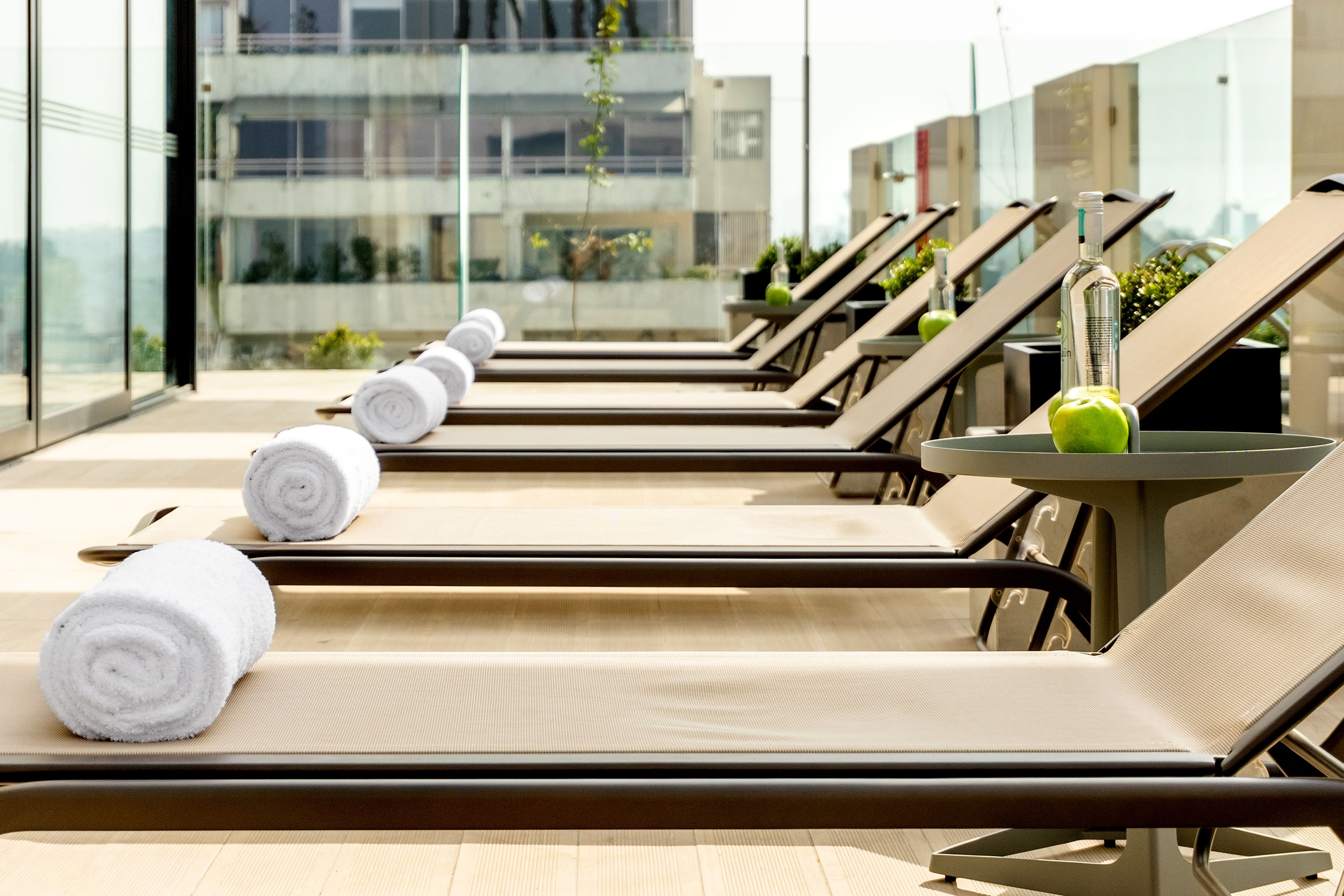 A row of sun loungers with towels, a table with a bottle and an apple, and a view of a building through large windows.