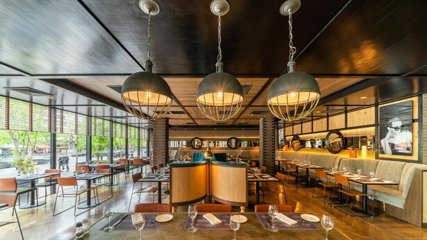 Un restaurante con una decoración moderna, que presenta una zona de bar central, varias mesas de comedor y lámparas colgantes.