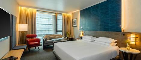 A hotel room with a large bed, a desk, a red chair, a sofa, a TV, and a window with curtains.