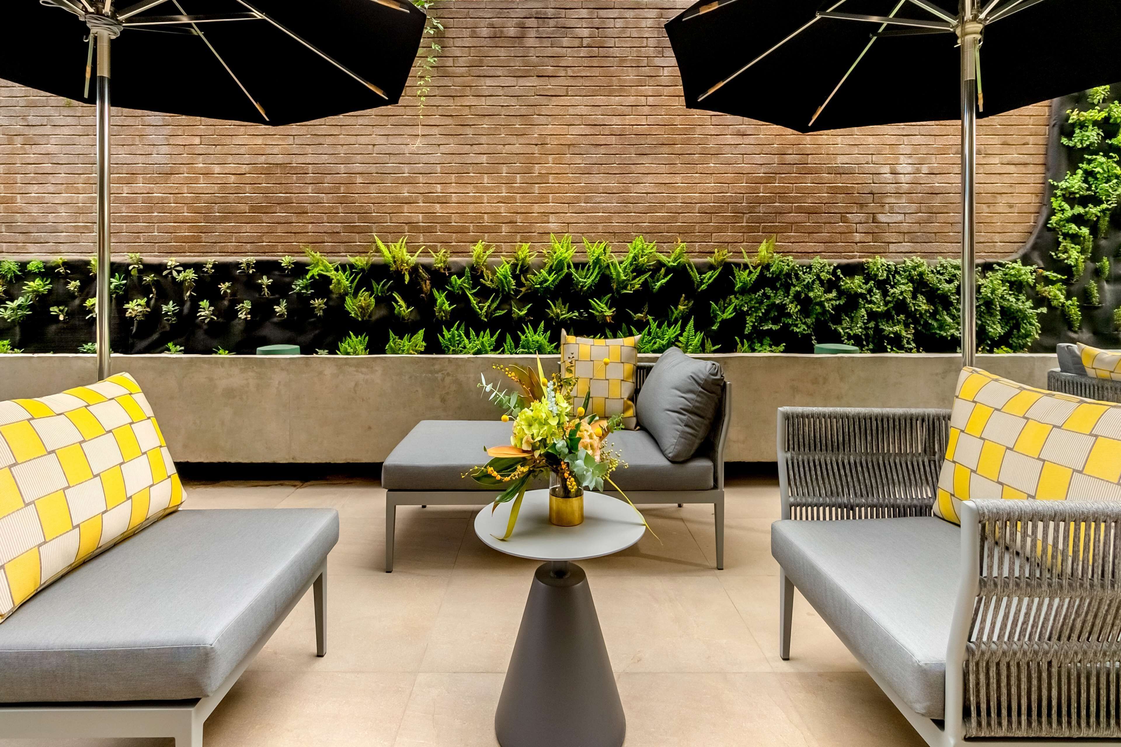 A modern outdoor seating area with grey sofas and yellow patterned cushions, a central round table with a vase of flowers, and black umbrellas providing shade.
