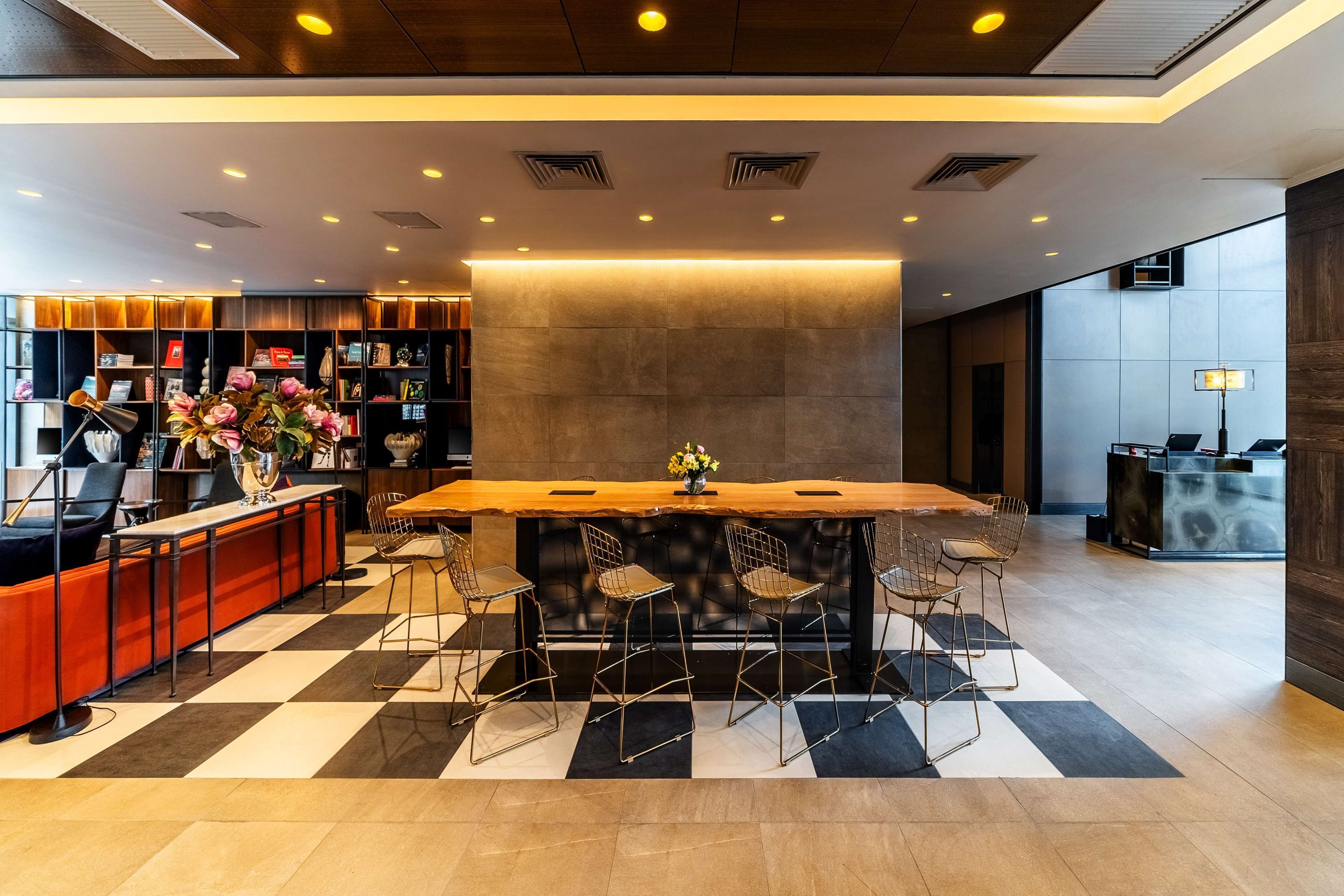 A modern hotel lobby with a checkered floor, a central bar area with stools, a bookshelf, and a reception desk.