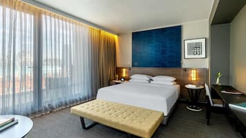 A hotel room with a large bed, a bench, a desk, and a view of the city through sheer curtains.