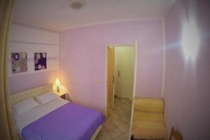 Ein kleiner, kompakter Raum mit einem Bett, einem Nachttisch und einer Tür zu einem weiteren Raum.