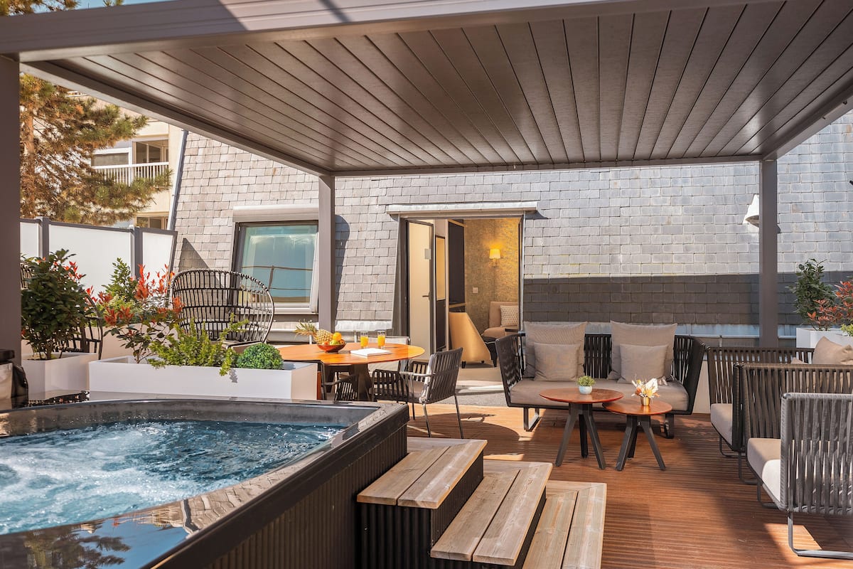 A covered outdoor patio with a hot tub, wooden furniture, and a brick wall.