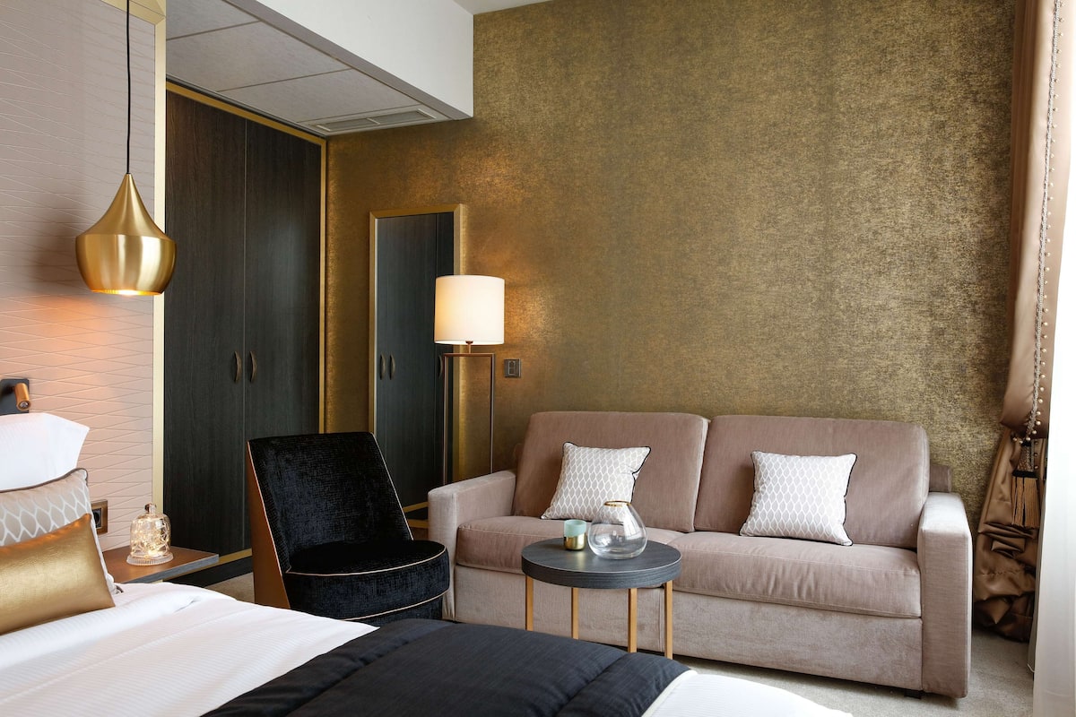 A hotel room with a bed, a sofa, a chair, a small round table, and a lamp.