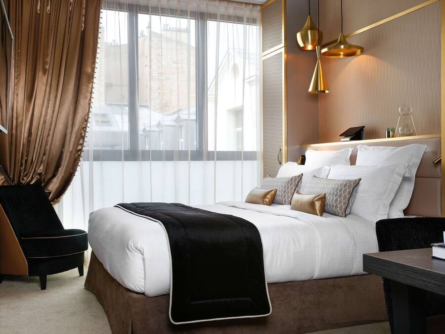 Een moderne hotelkamer met een groot bed, een zwarte fauteuil, een houten nachtkastje en een groot raam met dunne gordijnen.