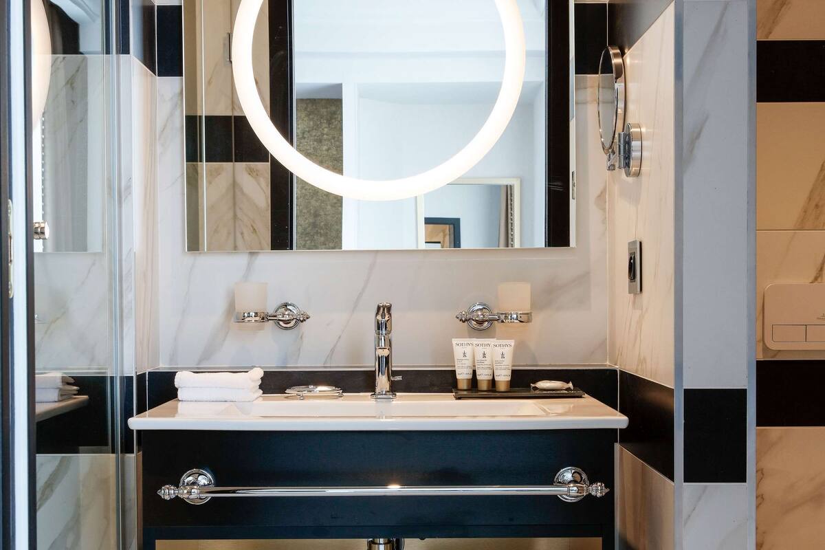 A modern bathroom with a black and white marble vanity, a glass shower enclosure, and a large mirror with a circular light.