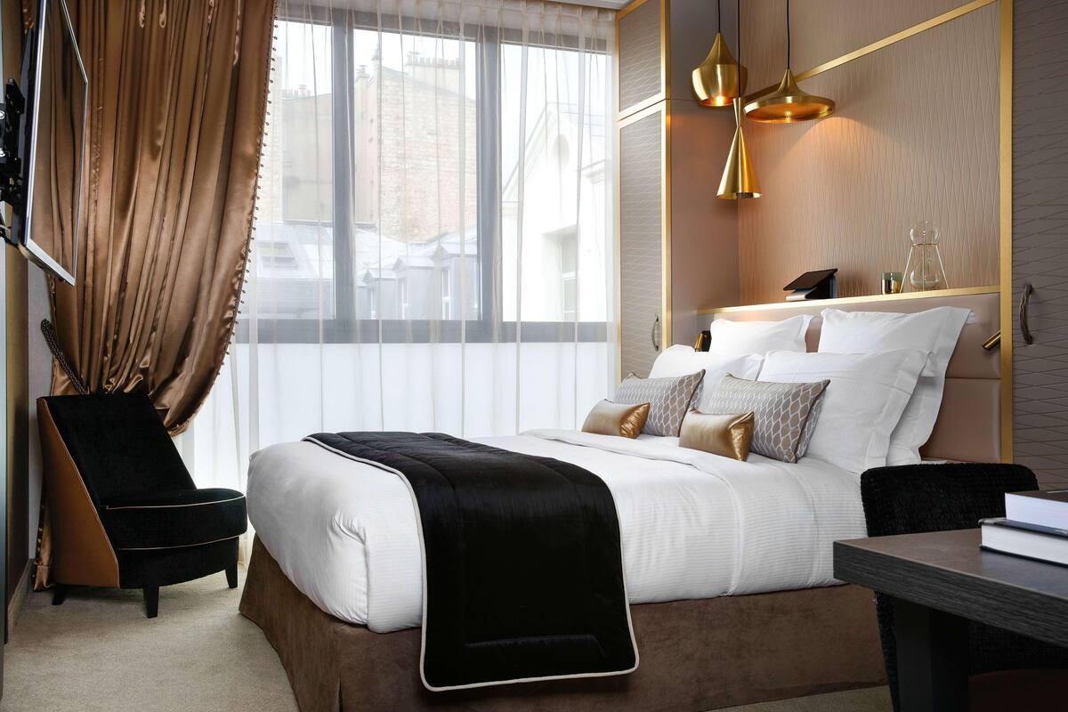 A hotel room with a bed, a chair, a desk, and a large window with curtains.