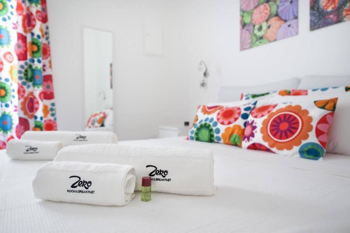 A hotel room with a neatly made bed, colorful floral pillows, and rolled white towels with the brand name 'Zero' visible.