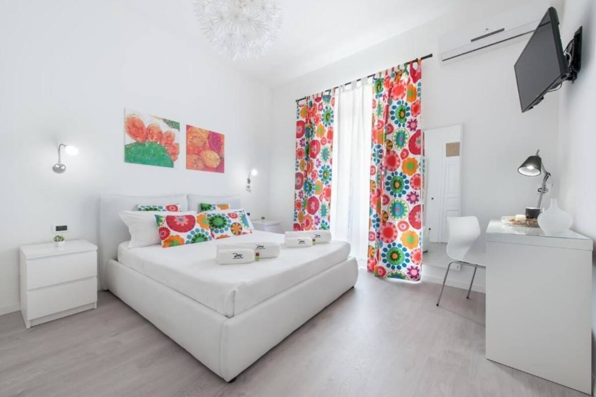 A modern bedroom with a white bed, colorful curtains, a desk, and a TV.