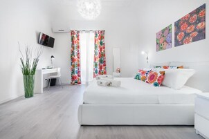 Una camera da letto moderna con un letto bianco, tende floreali colorate, una scrivania con una sedia e un televisore montato a parete.