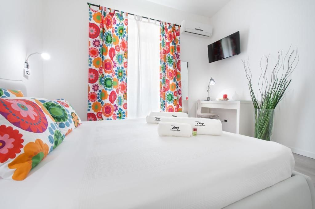 A hotel room with a bed, colorful curtains, a flat-screen TV, a bedside table with a lamp, and a vase with plants.