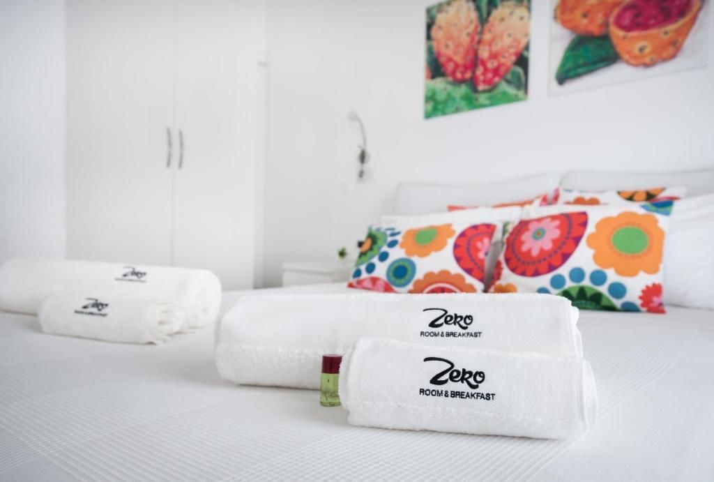 Un lit bien fait, avec une literie blanche et des coussins aux motifs floraux colorés, une serviette blanche ornée du logo de l’hôtel et une armoire blanche dans une chambre d’hôtel.