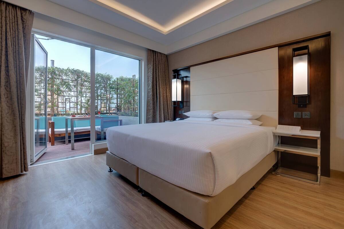 A modern hotel room with a large bed, a bedside table, a built-in TV, and a sliding glass door leading to a pool area.
