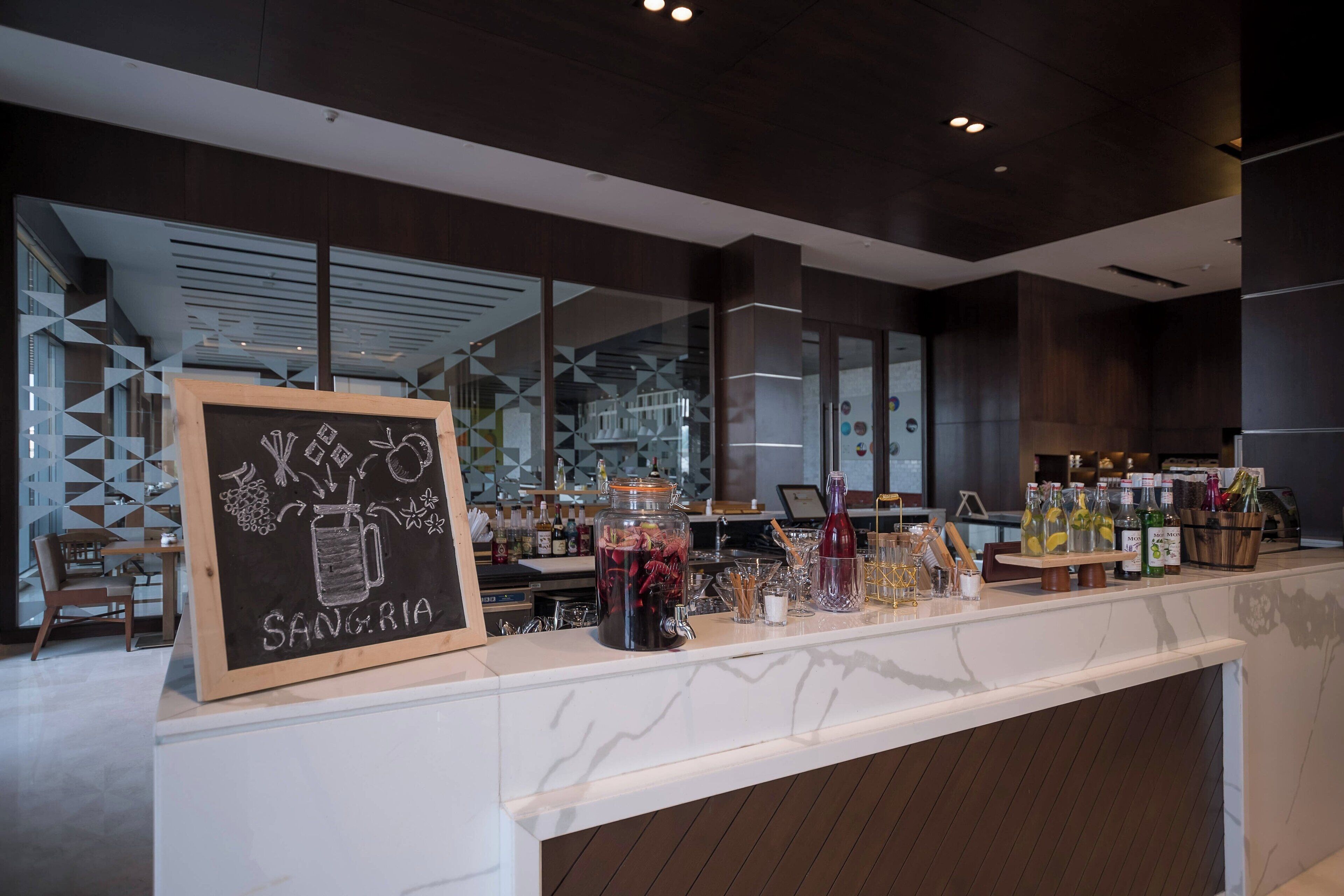 A bar area with a chalkboard sign reading "SANGRIA," a variety of bottles and glasses on the counter, and a seating area with a geometric wall design in the background.