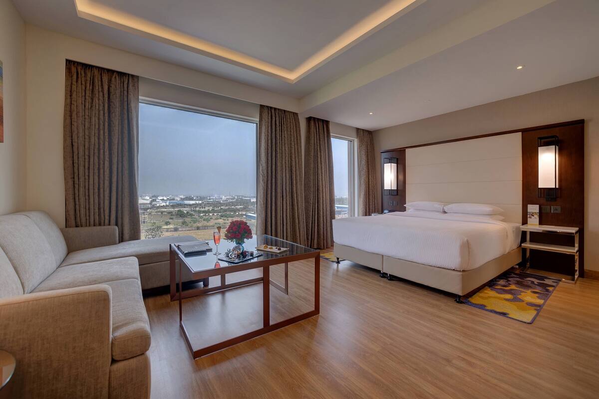 A hotel room with a bed, a sofa, a glass coffee table, and large windows offering a city view.