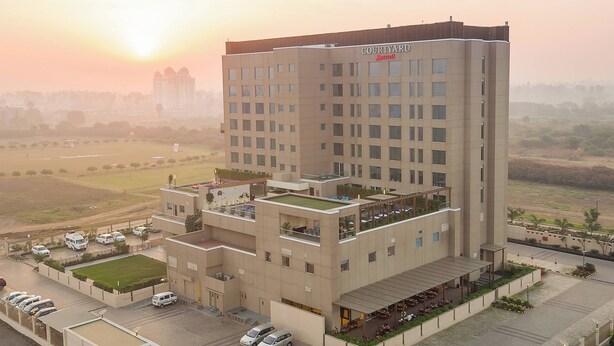 A multi-story hotel with a flat roof terrace, a parking area, and a view of the surrounding landscape. - Courtyard by Marriott Surat (Surat)