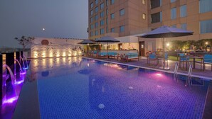 A hotel pool area with a tiled pool, lounge chairs, umbrellas, and a building with multiple windows. - Courtyard by Marriott Surat (Surat)