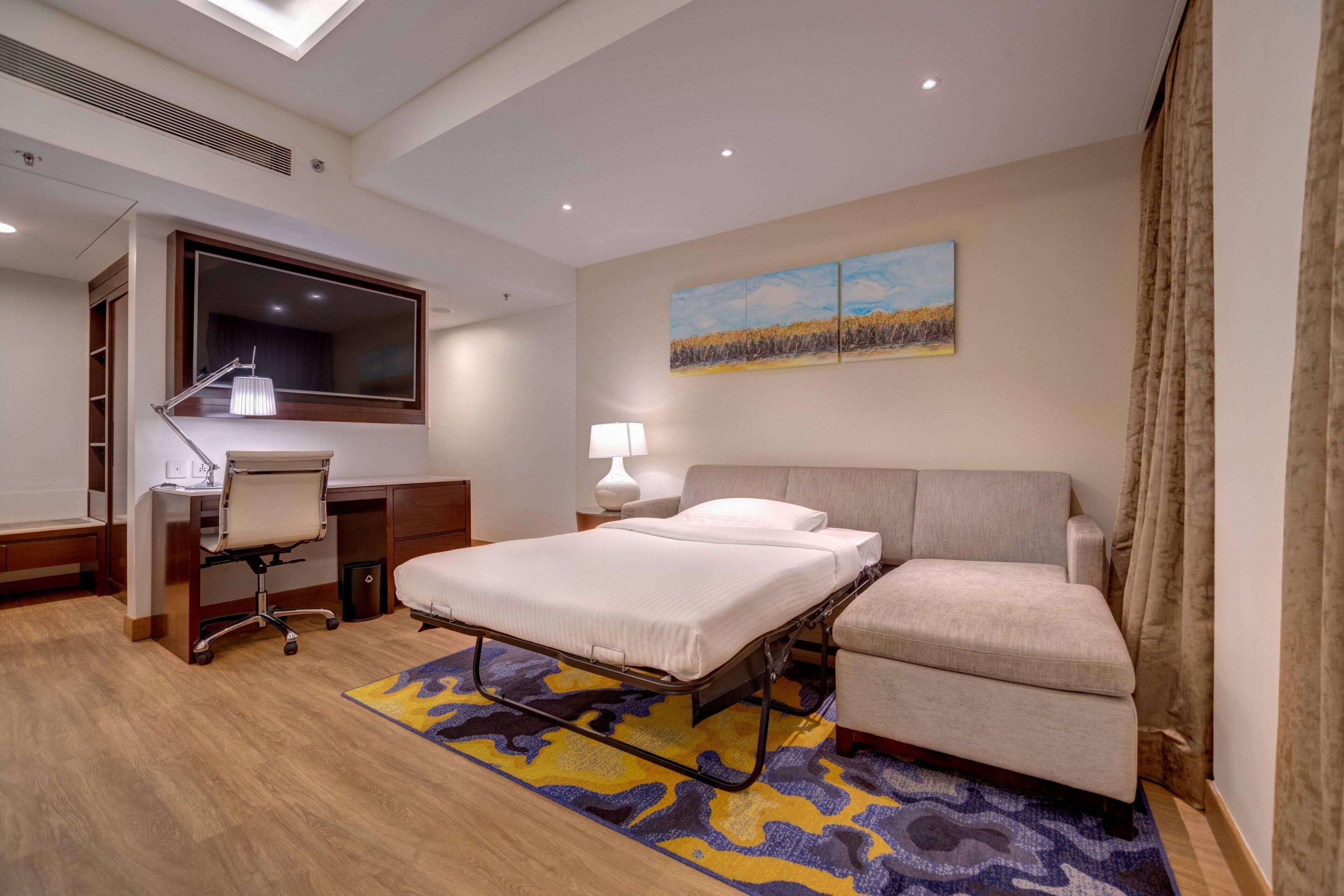 Una habitación de hotel con cama, escritorio, silla, lámpara y un cuadro en la pared.