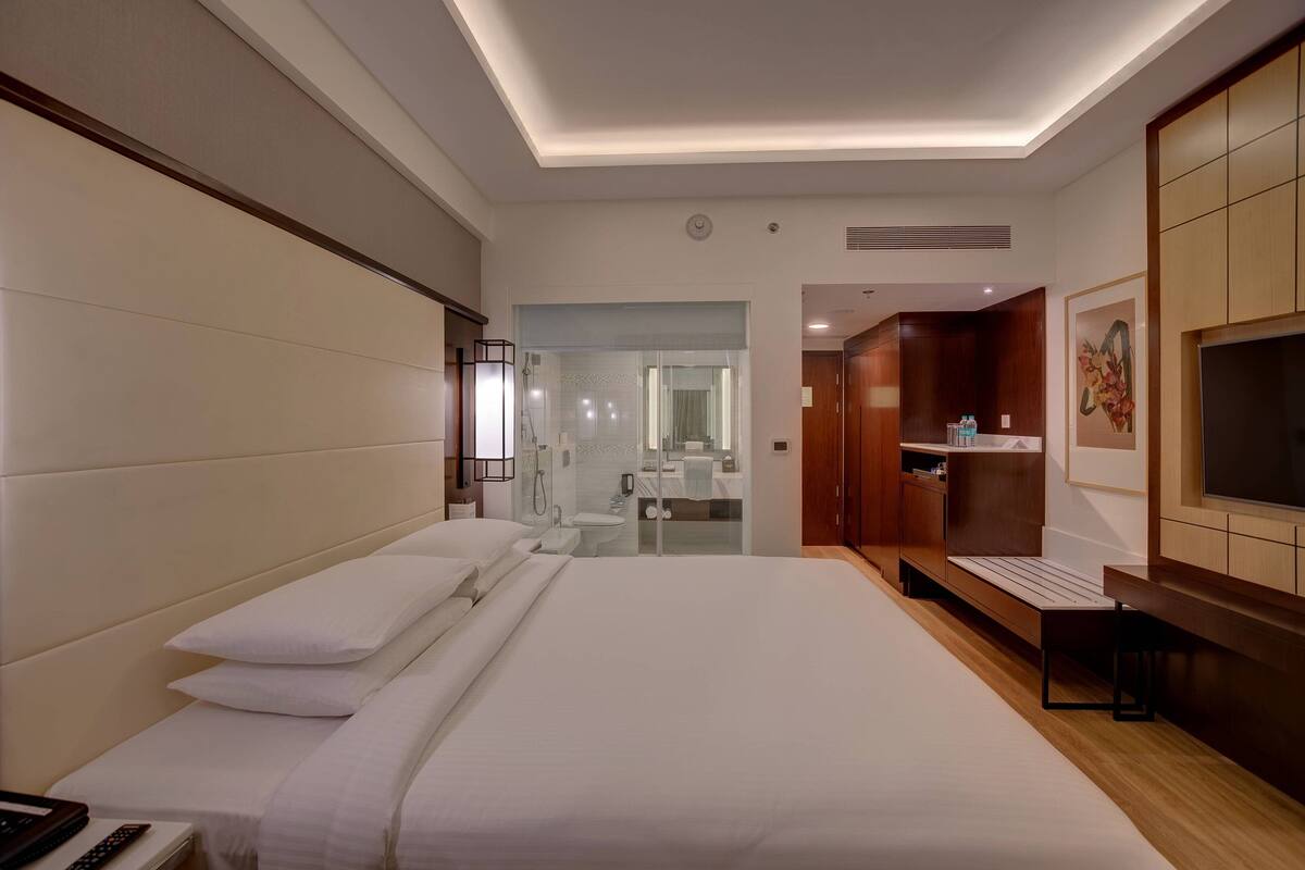 A hotel room with a large bed, a TV, a desk with a chair, and a bathroom visible through a glass door.