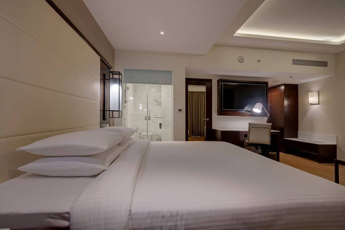 A hotel room with a large bed, a desk, a chair, a mirror, and a shower area.