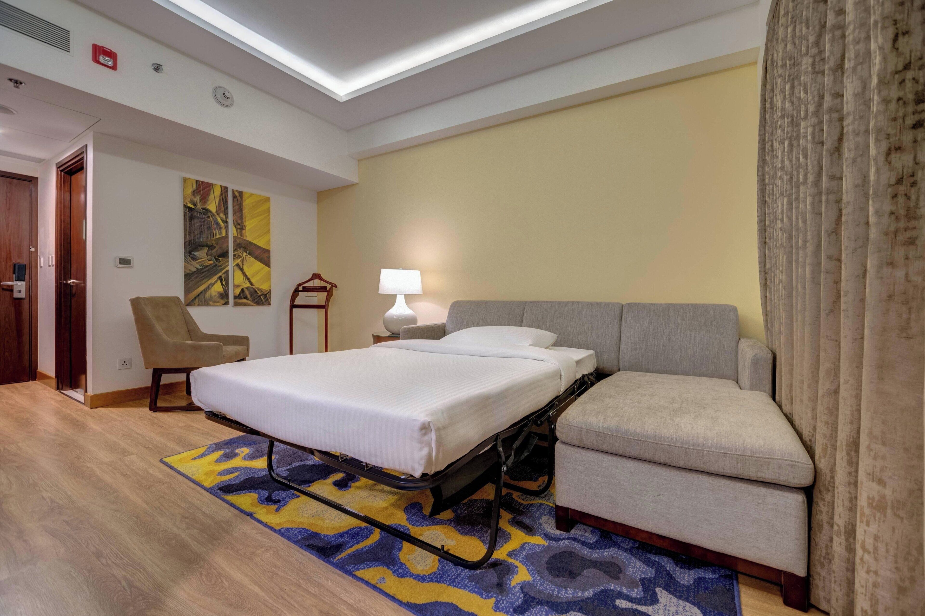 Habitación de hotel con cama, sofá, sillón, lámpara y un cuadro en la pared.