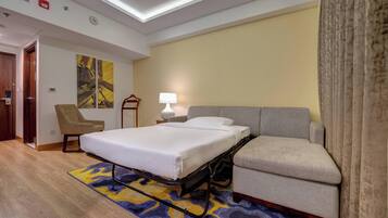 Habitación de hotel con cama, sofá, sillón, lámpara y un cuadro en la pared.