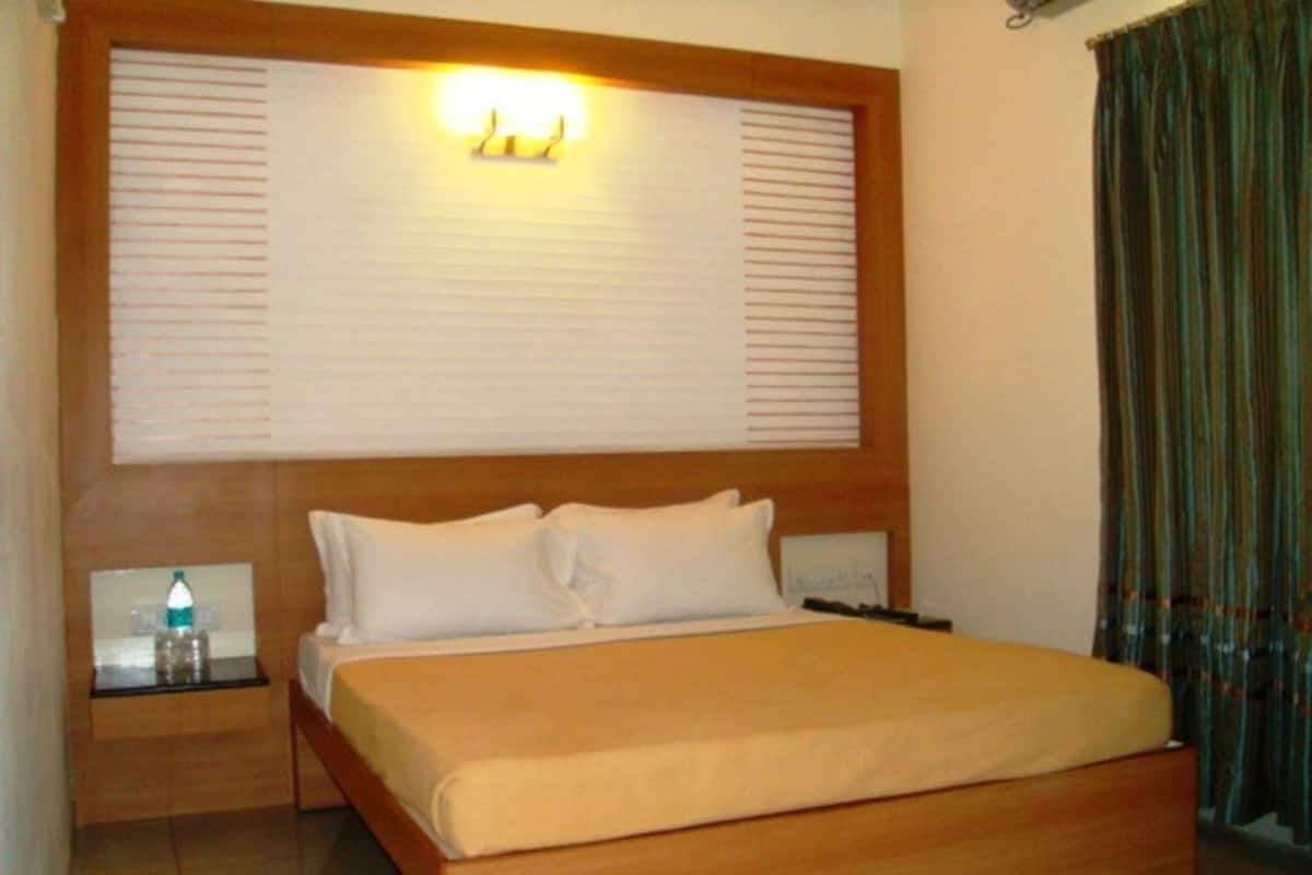 Quarto de hotel com cama grande, cabeceira de madeira, mesas de cabeceira com luminárias e cortina na janela.