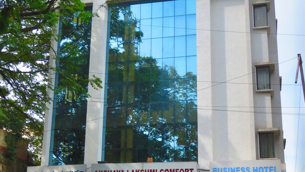 En bygning med flere etager, et skilt med "Akshaya Lakshmi Comfort Business Hotel" og en trappe, der fører til indgangen.