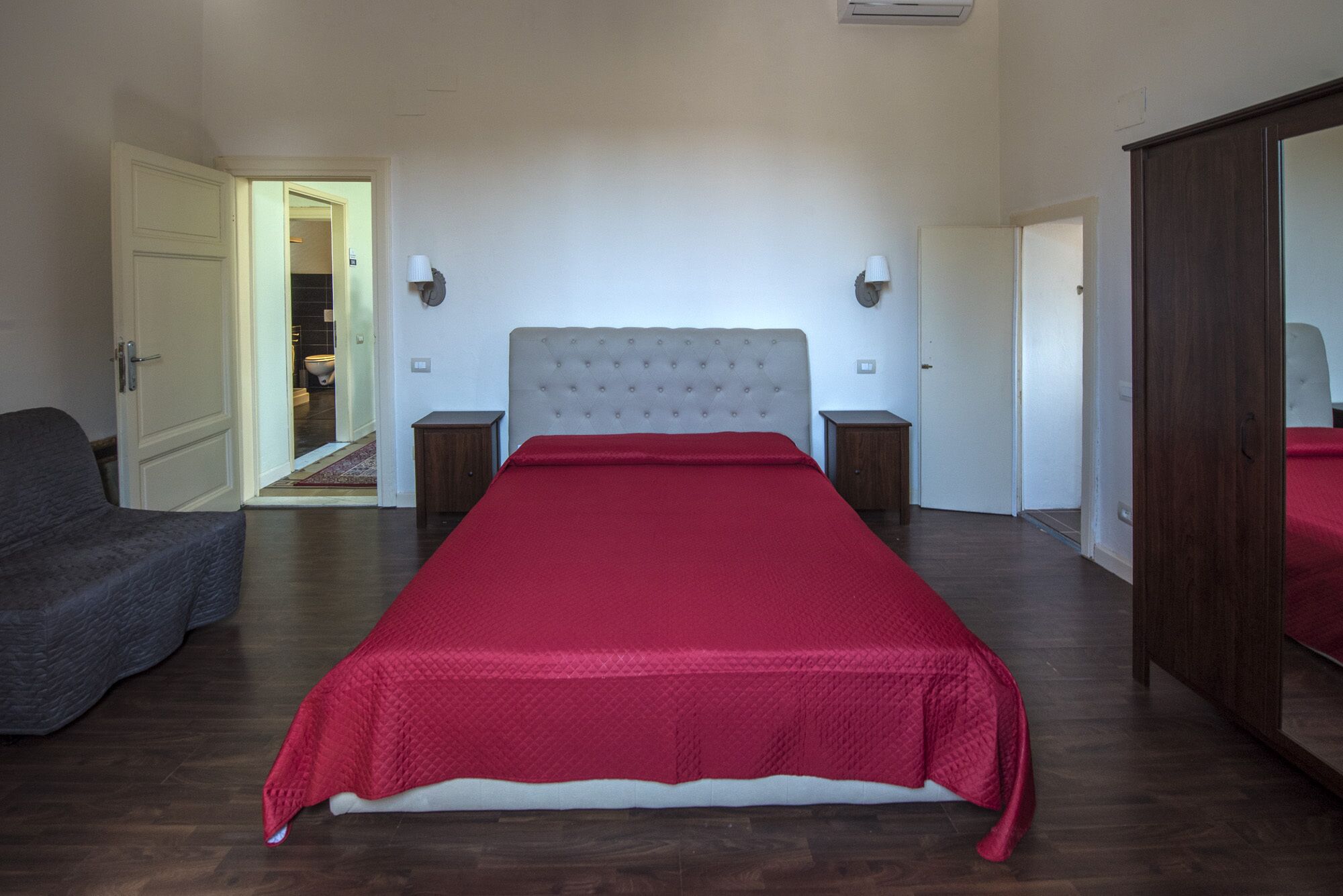 A bedroom with a large bed, a wardrobe, a bedside table, a lamp, and a door leading to another room.