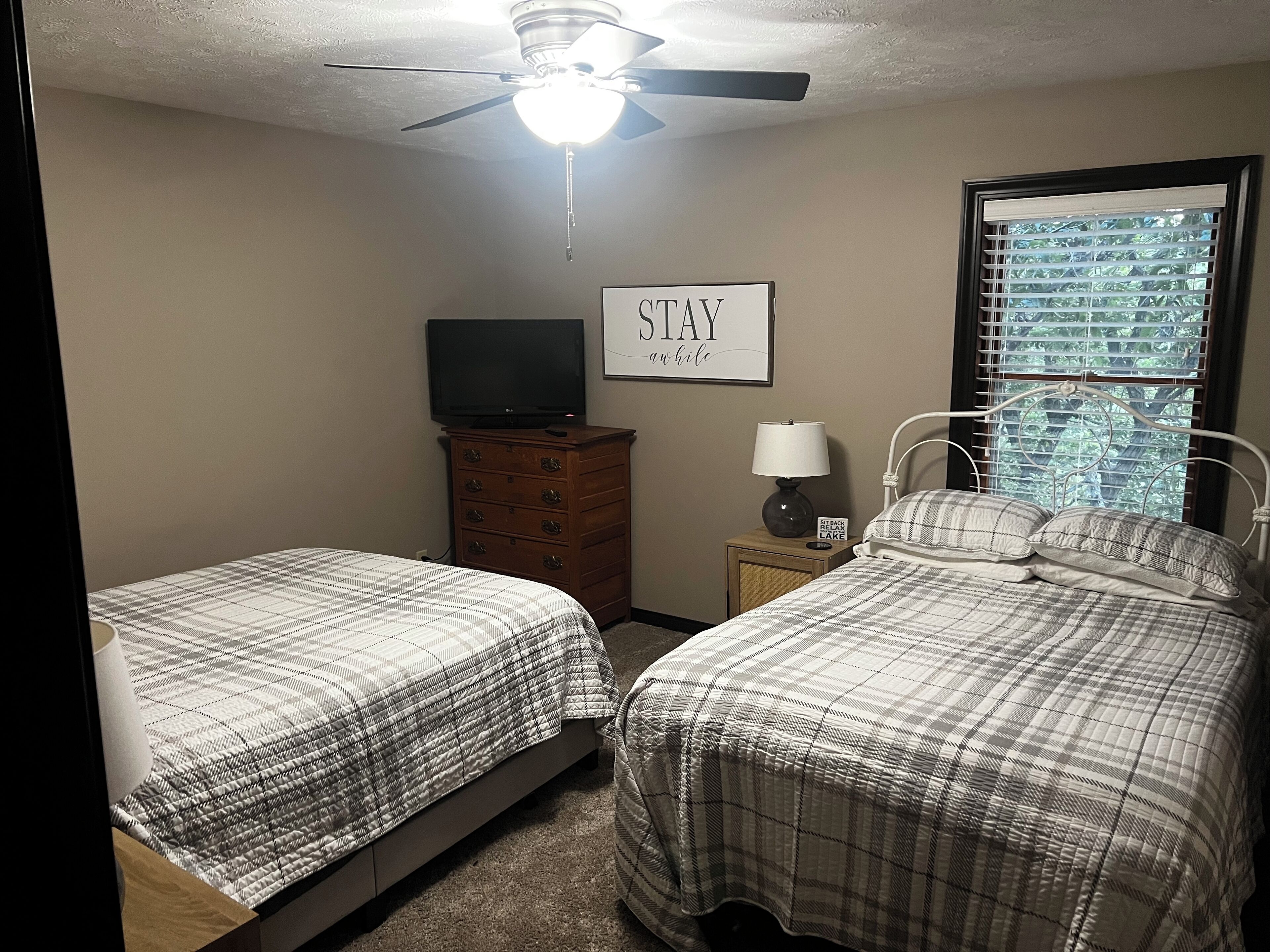 A bedroom with two beds, a ceiling fan, a TV on a dresser, and a window with blinds.