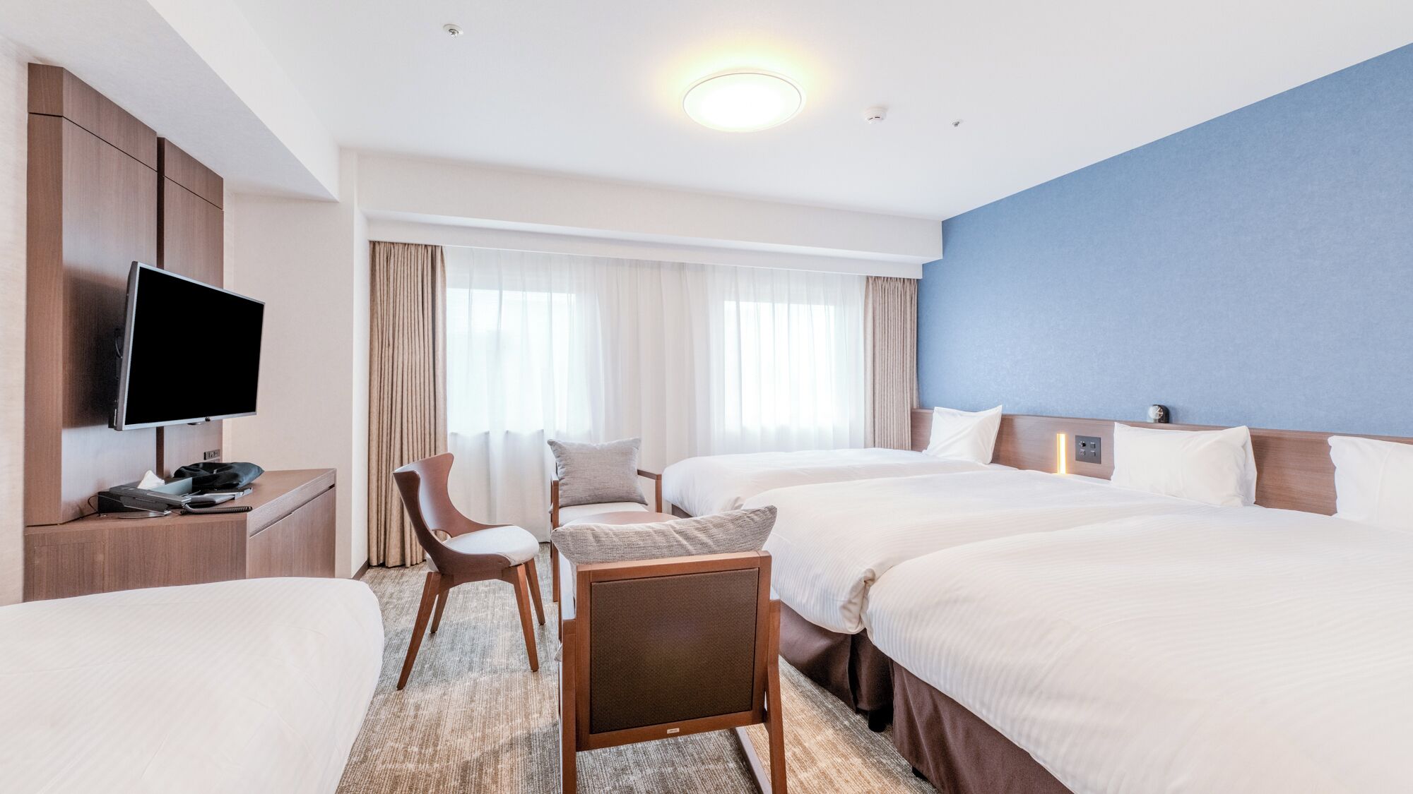 A hotel room with a flat-screen TV, two beds, a desk with a chair, and a large window with curtains.