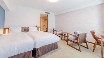 Quarto de hotel com uma cama grande, duas cadeiras, uma mesinha e um banheiro visível através de uma porta aberta.