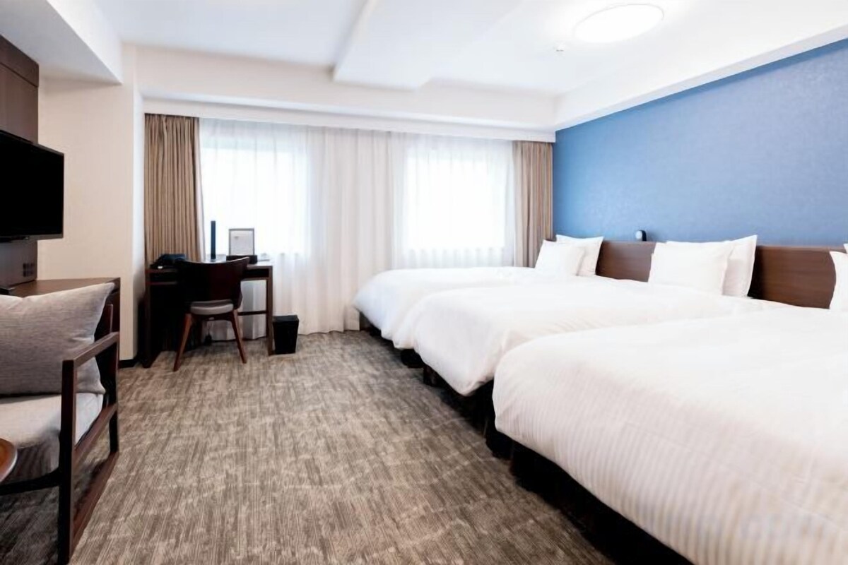 A hotel room with a large bed, a desk, a chair, a television, and a window with curtains.