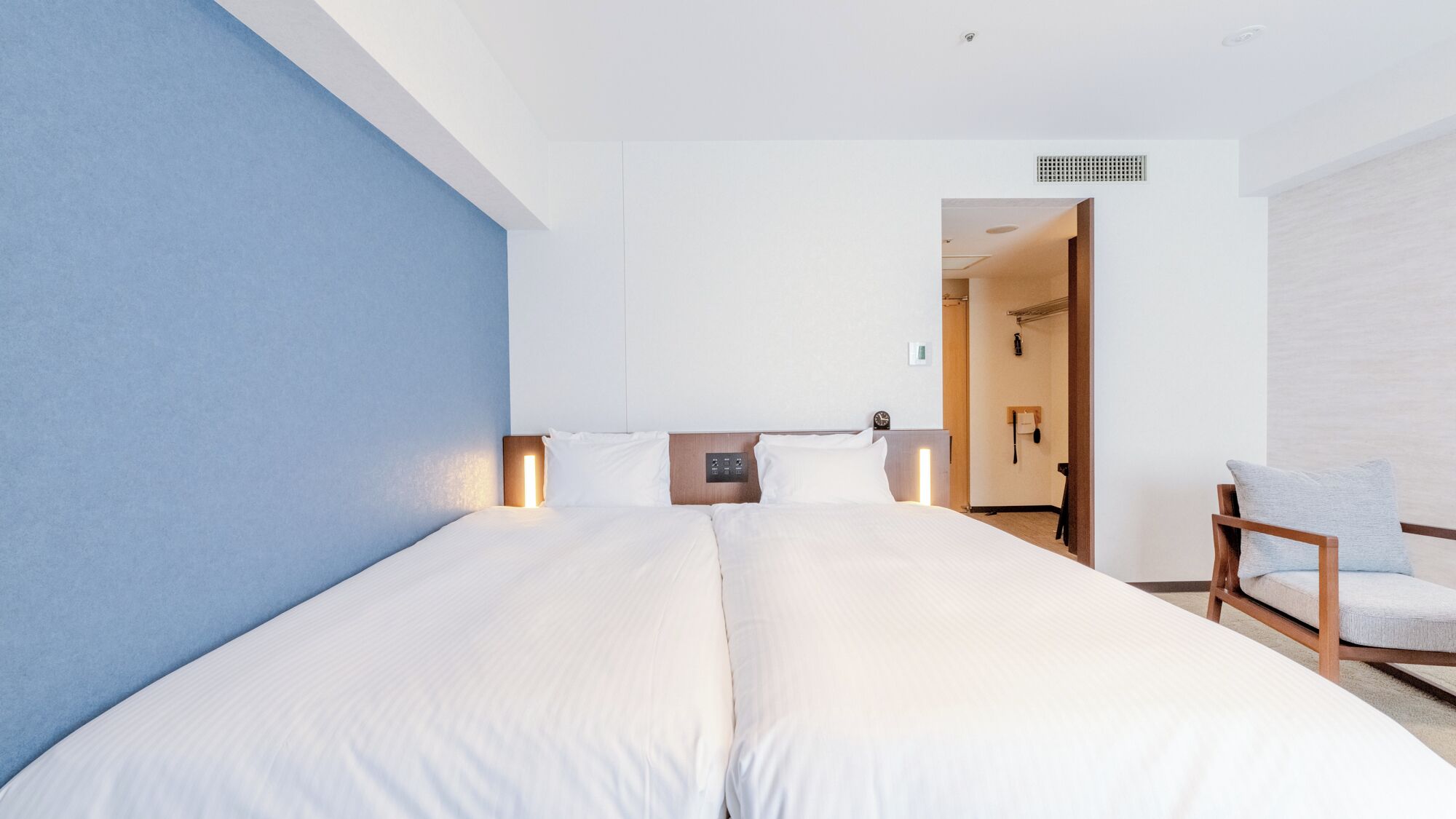 Quarto de hotel moderno com uma cama grande, abajures, uma cabeceira de madeira e uma cadeira.