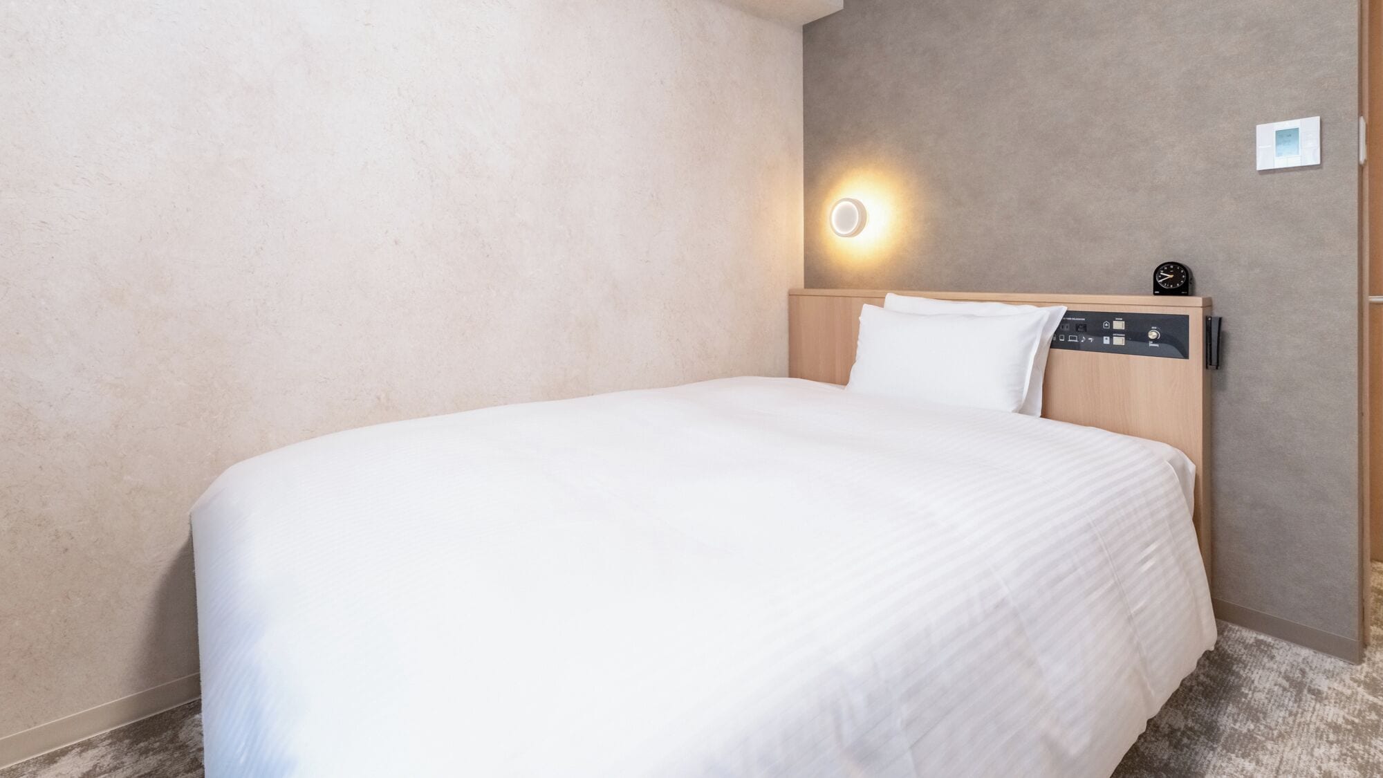 Habitación de hotel con cama, cabecera con panel de control, luz empotrada en la pared y termostato.