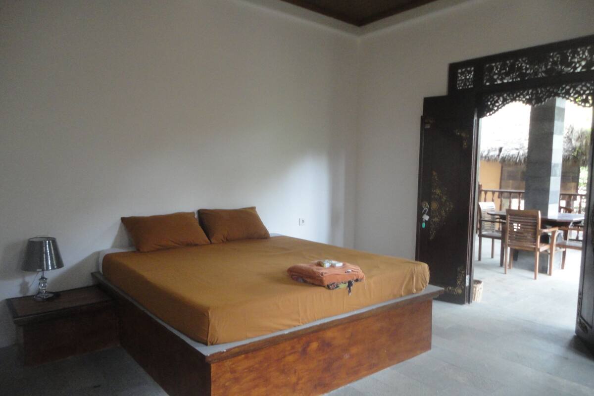 A bedroom with a large bed, a nightstand with a lamp, and a view of an outdoor patio with chairs and a table.