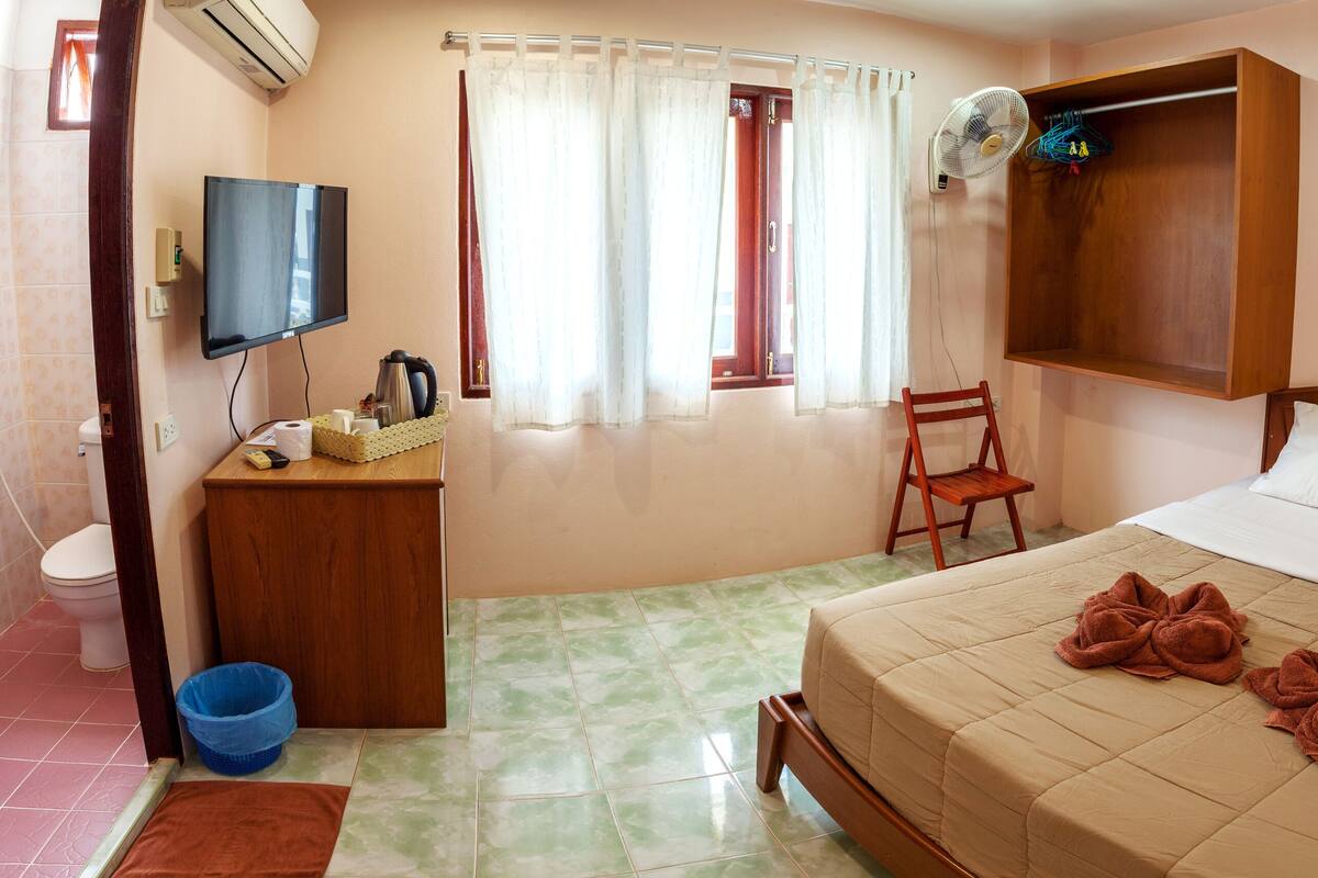 A hotel room with a bed, a wooden nightstand, a television, a window with curtains, and a fan.