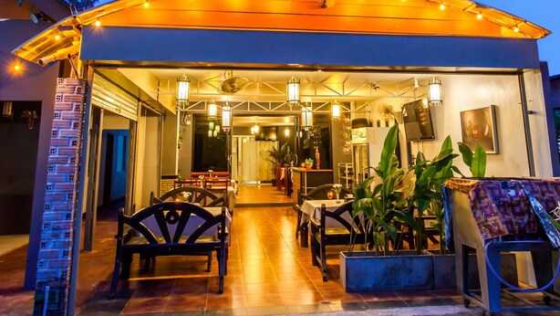 Ein Restaurant mit Außenbestuhlung, einer überdachten Terrasse und Blick auf ein hohes Gebäude im Hintergrund.