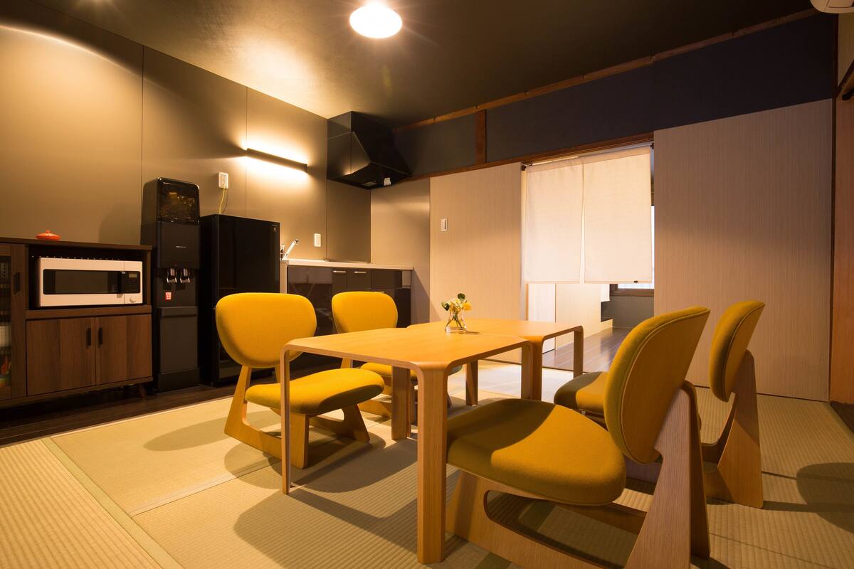 A modern kitchen with a microwave, refrigerator, and wooden cabinets. There is a dining table with yellow chairs and a small vase with flowers.