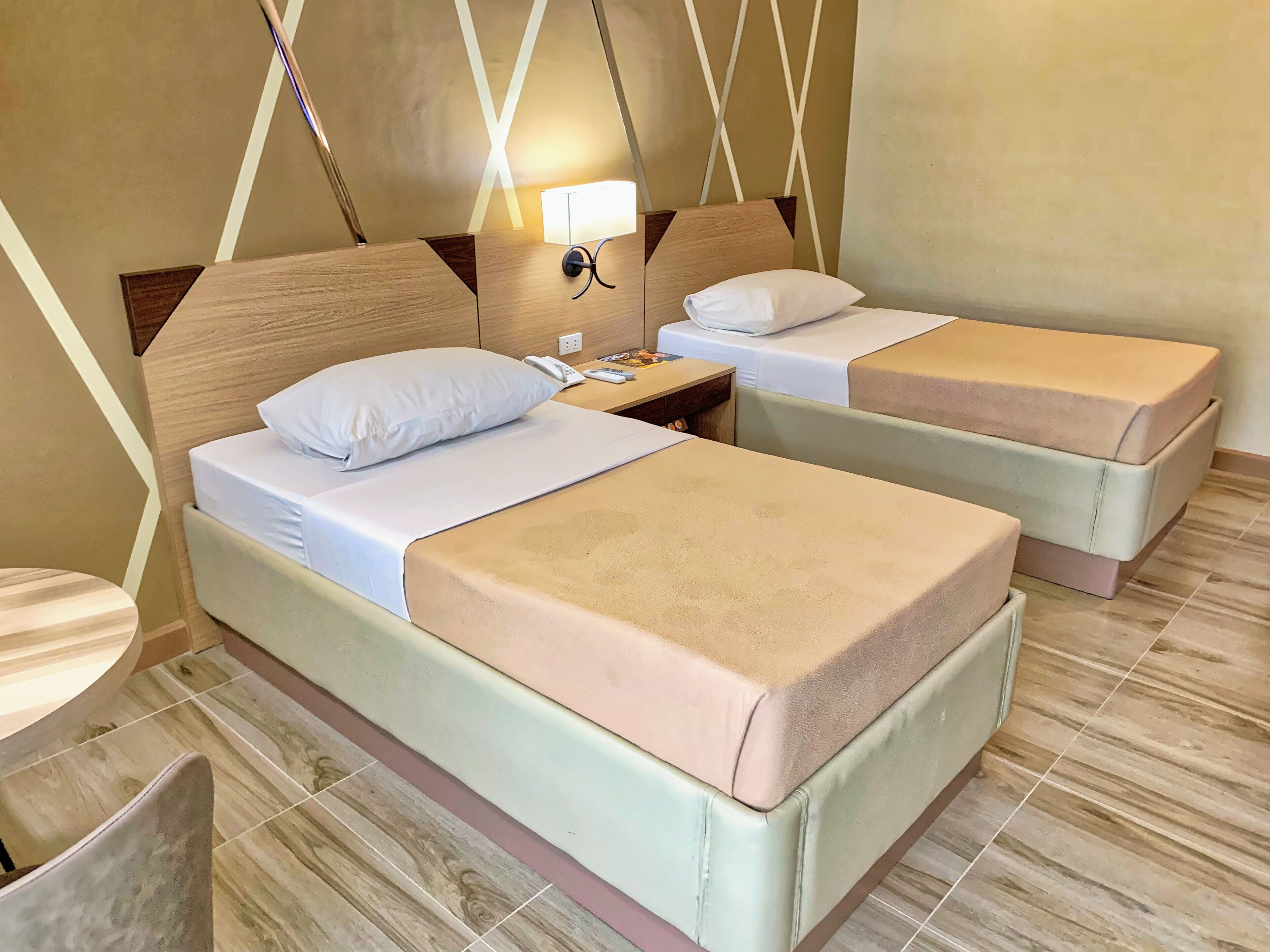 Habitación de hotel con dos camas, cada una con almohada y manta. Se observa una mesita de noche con lámpara y teléfono. La habitación tiene cabecero de madera y suelo de baldosas.