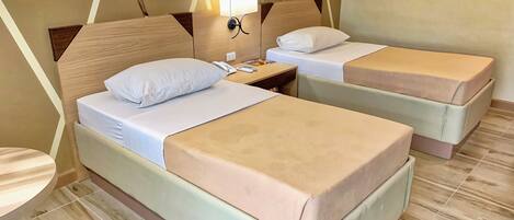 Habitación de hotel con dos camas, cada una con almohada y manta. Se observa una mesita de noche con lámpara y teléfono. La habitación tiene cabecero de madera y suelo de baldosas.