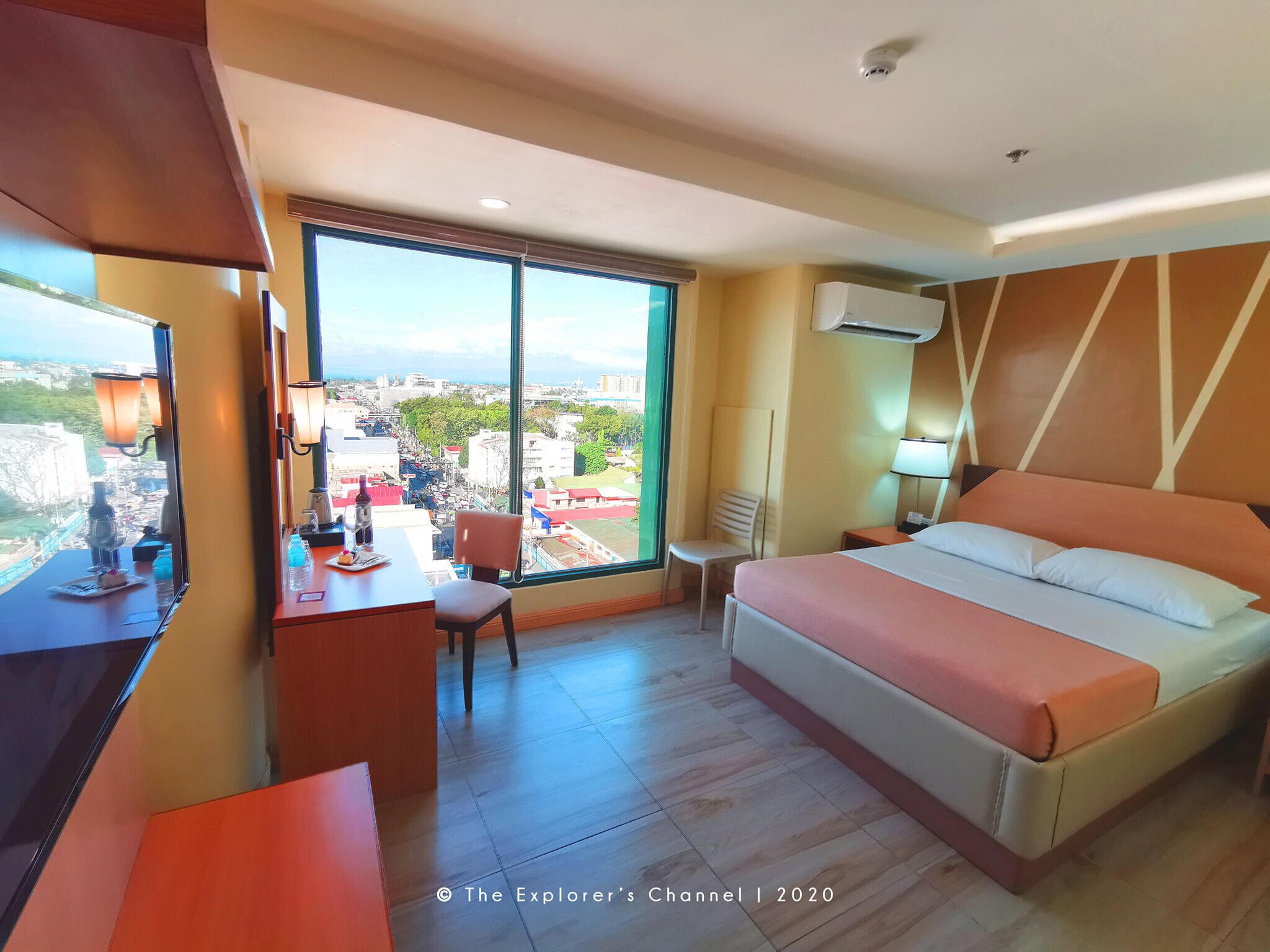 A hotel room with a bed, desk, chair, and a large window offering a city view.