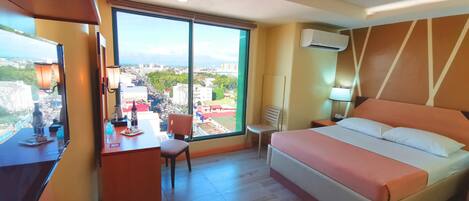 A hotel room with a bed, desk, chair, and a large window offering a city view.