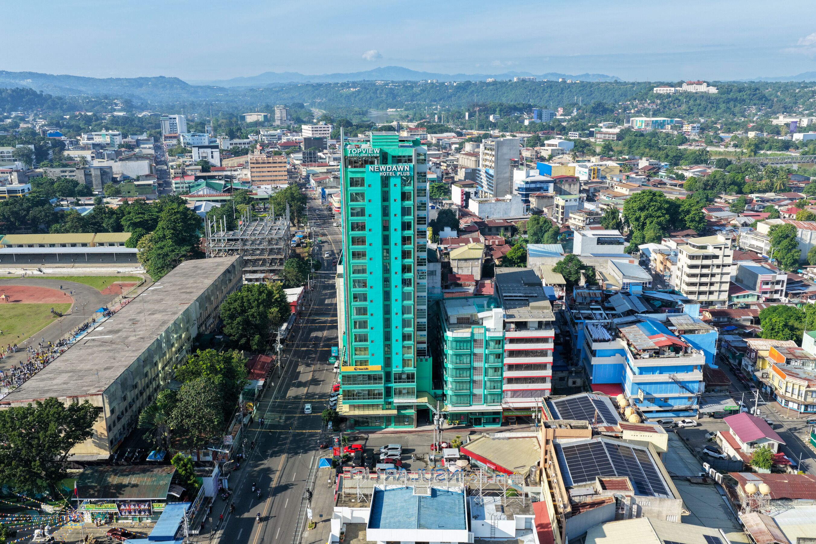 A high-rise hotel with multiple floors, surrounded by a dense urban area with various buildings and a sports field.
