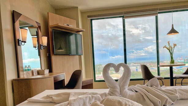 A hotel room with a bed, a desk, a chair, a TV, a window with a city view, and towels arranged in a heart shape. - New Dawn Hotel Plus (Cagayan de Oro)